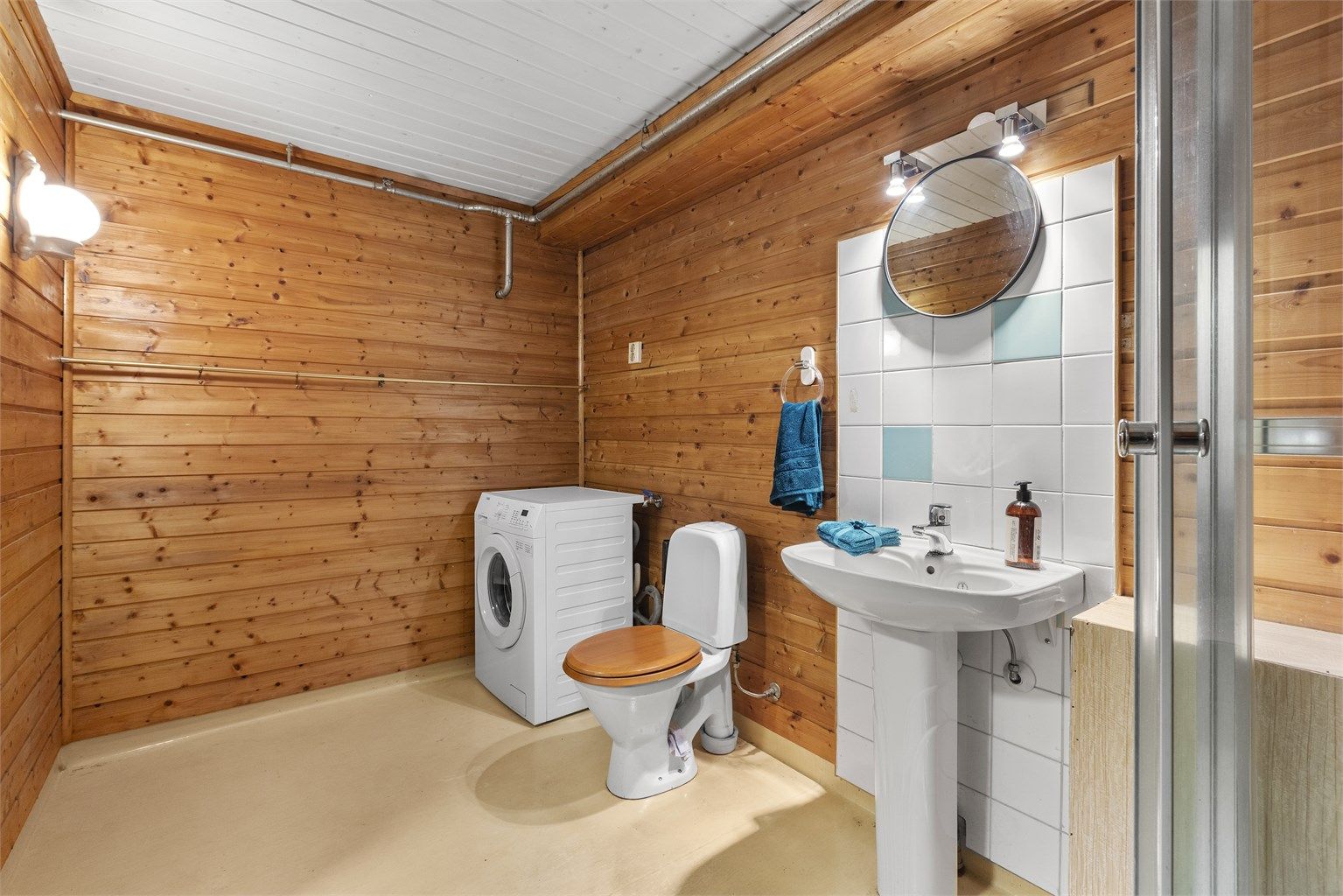 Badet er fra byggeår med gulvbelegg og panel på vegger og i himling. Rommet er innredet med servant, dusjkabinett og toalett, med opplegg for vaskemaskin.