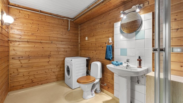 Badet er fra byggeår med gulvbelegg og panel på vegger og i himling. Rommet er innredet med servant, dusjkabinett og toalett, med opplegg for vaskemaskin.