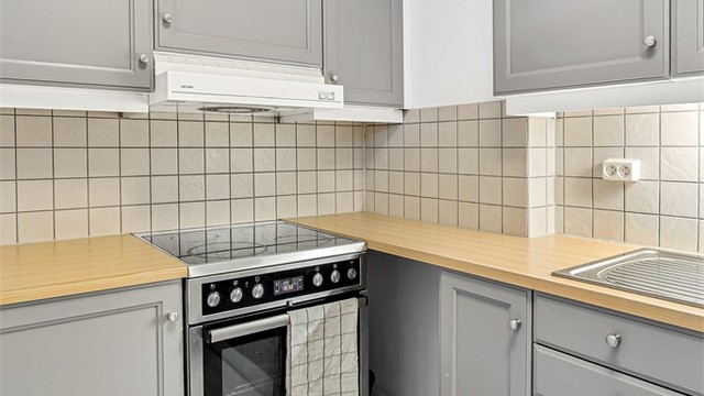 Kjøkkeninnredningen har profilerte fronter i grå utførelse. Videre er benkeplaten av laminat og det er fliser på vegg mellom benkeplaten og overskap.
