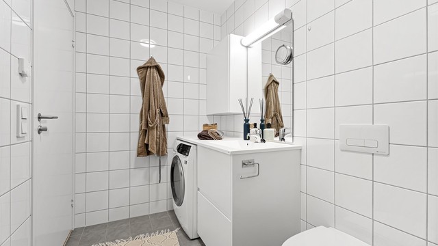Videre er det vegghengt toalett, servant med plassbygd nisje til vaskemaskin, arbeidsbenk samt godt med lagringsplass i skuffer og overskap.
