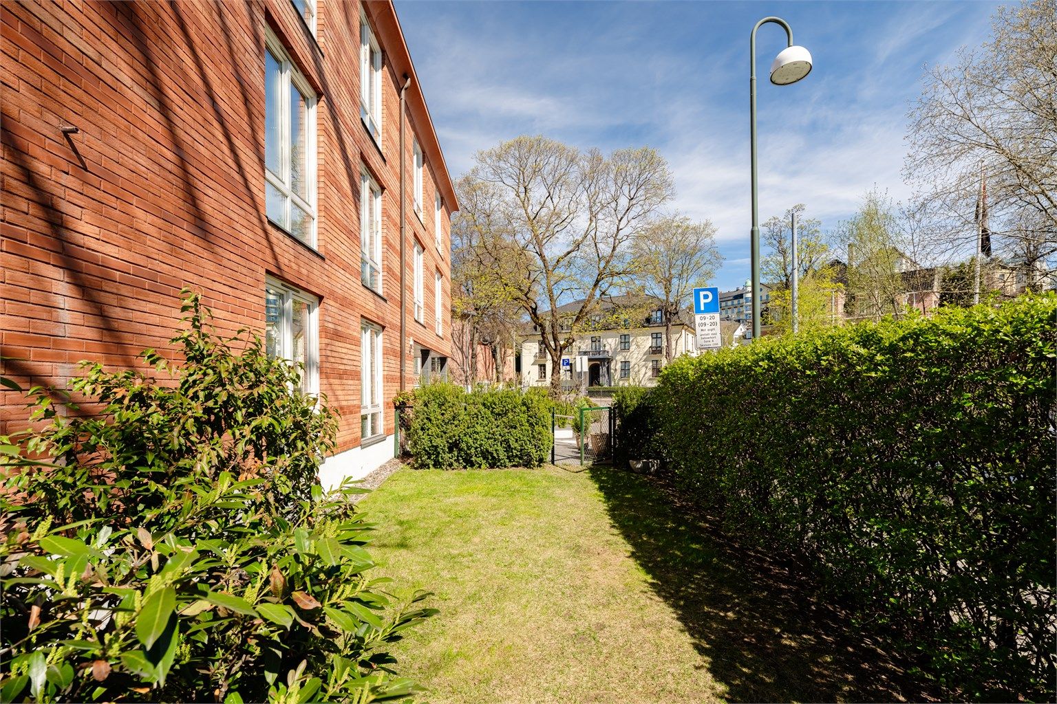 Pent opparbeidet plen med busker og blomster rundt bygningen. Hellelagt inngangsparti med bred trapp og asfaltert nedkjørsel til garasjeanlegg. 