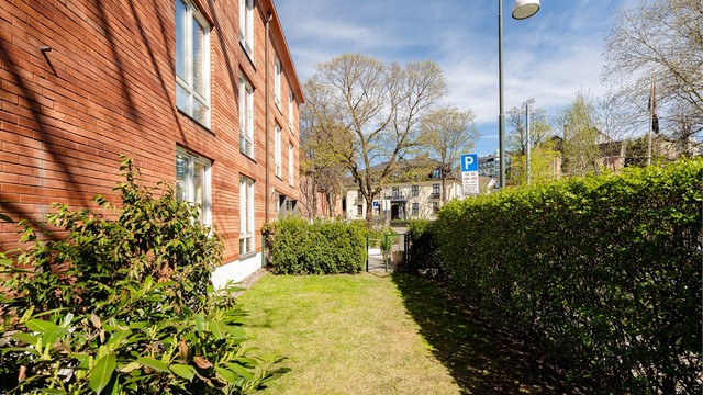 Pent opparbeidet plen med busker og blomster rundt bygningen. Hellelagt inngangsparti med bred trapp og asfaltert nedkjørsel til garasjeanlegg.