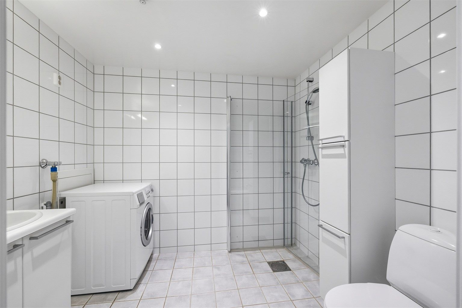 Flislagt bad med varmekabler i gulv og opplegg for vaskemaskin. Badet er innredet med dusjhjørnet med dører og vegghengt toalett. 