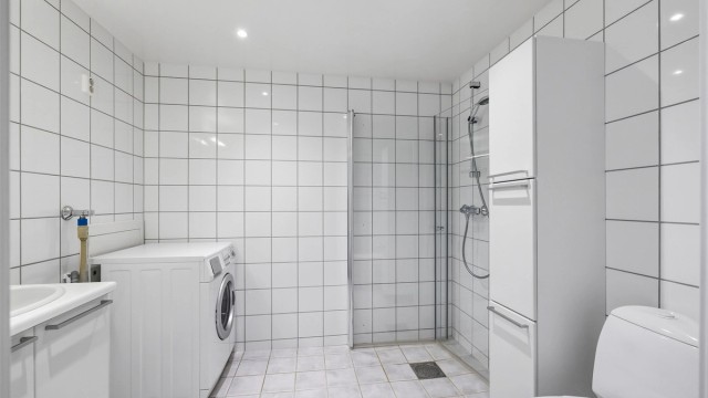 Flislagt bad med varmekabler i gulv og opplegg for vaskemaskin. Badet er innredet med dusjhjørnet med dører og vegghengt toalett.