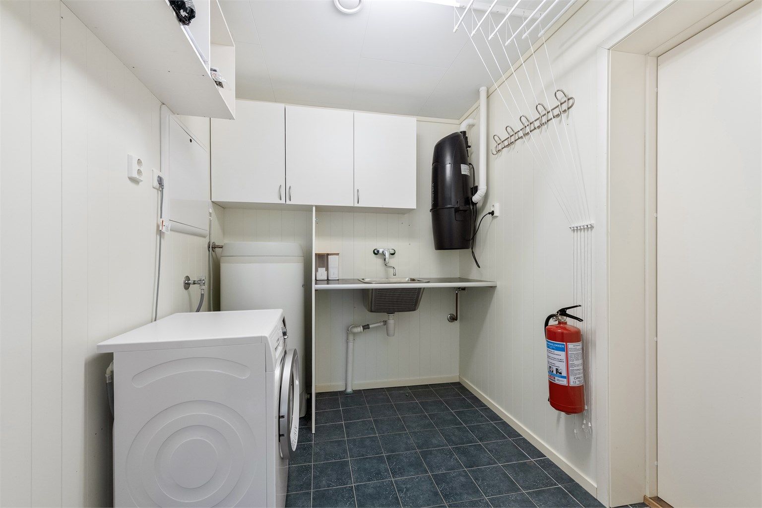 Praktisk vaskerom med flislagt gulv, utslagsvask og opplegg for vaskemaskin. Varmtvannsberederen er på 200L fra 2010