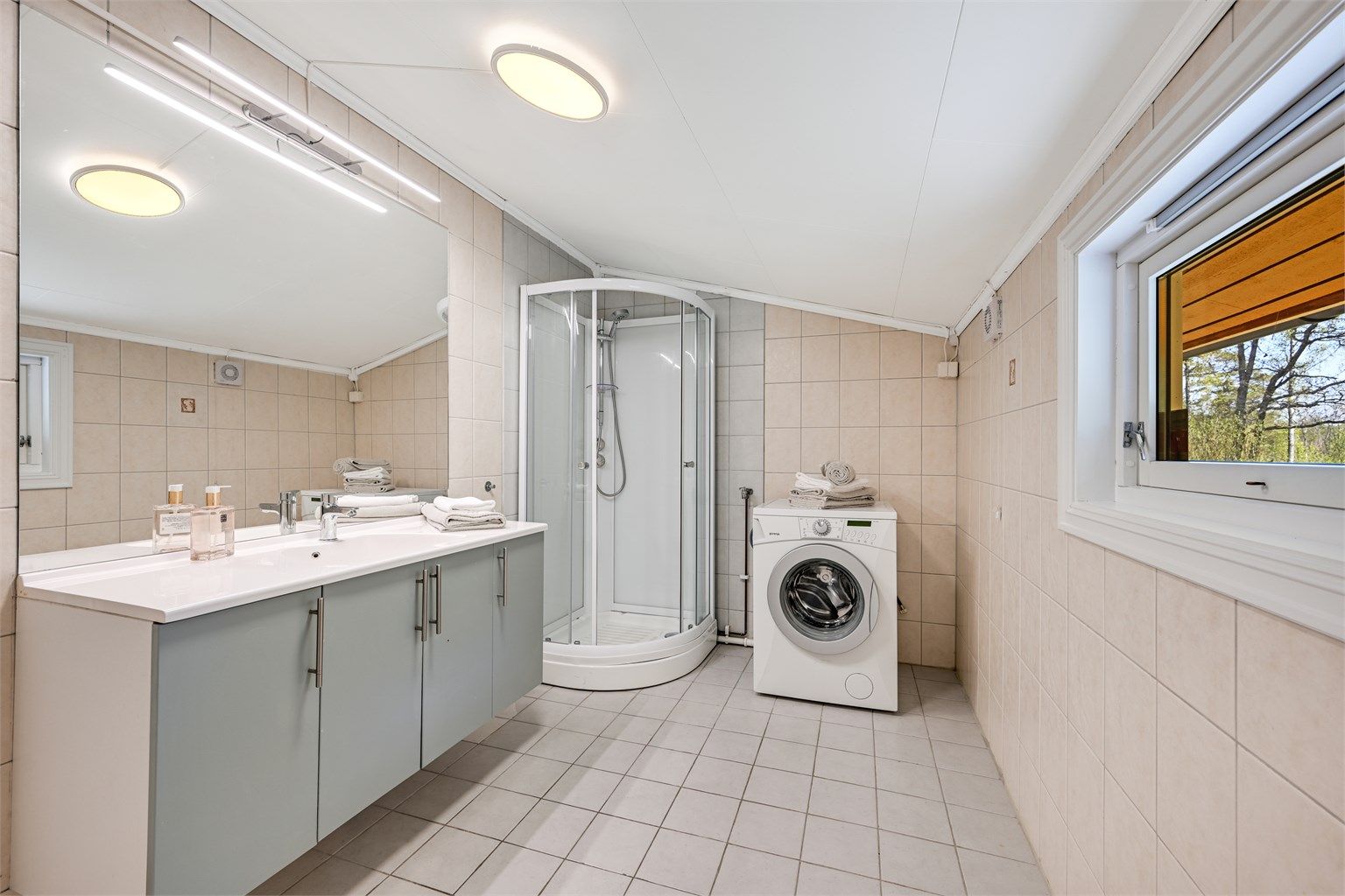Badet har flislagte gulv og vegger og er innredet med dusjkabinett, servant i baderomsinnredningen, samt opplegg for vaskemaskin