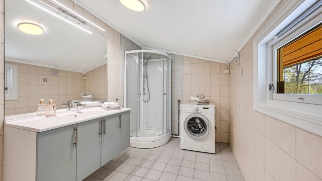 Badet har flislagte gulv og vegger og er innredet med dusjkabinett, servant i baderomsinnredningen, samt opplegg for vaskemaskin