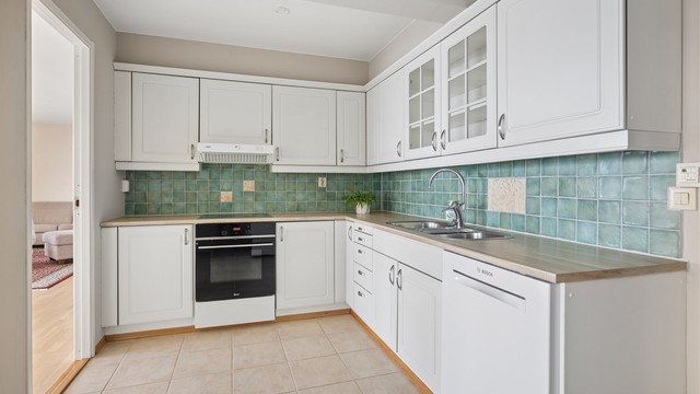 Kjøkkeninnredningen fremstår både pen og funksjonell, med rikelig benkeplass og gode oppbevaringsmuligheter i over- og underskap. De profilerte frontene gir et klassisk preg, mens glassfronter enkelte steder tilfører en elegant visuell variasjon.