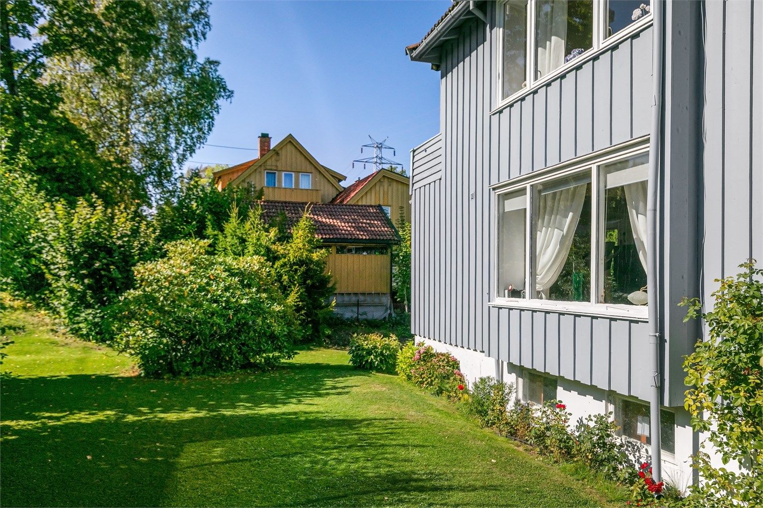 Fasaden er kledd med stående trekledning, og tomten er pent opparbeidet med plen og bed.