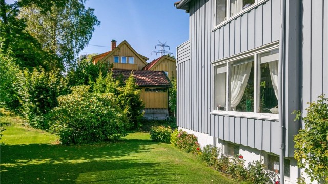 Fasaden er kledd med stående trekledning, og tomten er pent opparbeidet med plen og bed.