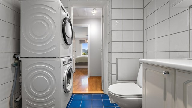 Badet har toalett, vegghengt servantinnredning, speil med overlys, dusjhjørne med dører og opplegg for vaskemaskin