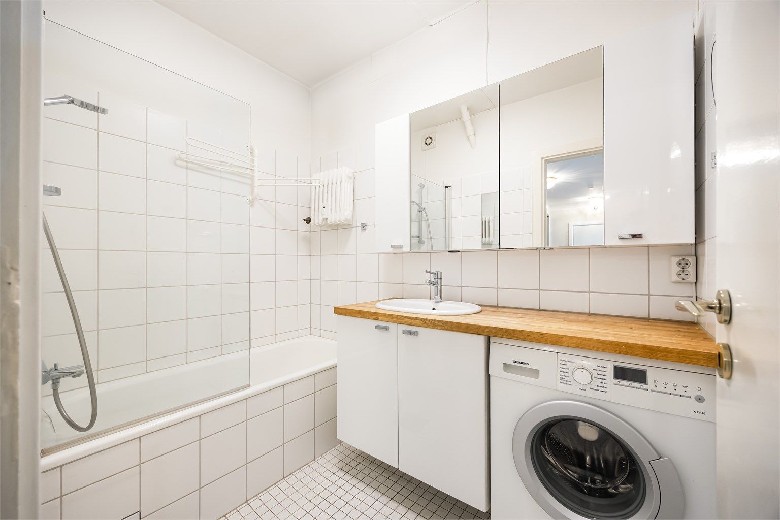 Lyst bad som er innredet med vegghengt servant med benkeplate i heltre og nedfelt vask. Innbygget badekar med glassvegg og dusjarmatur.