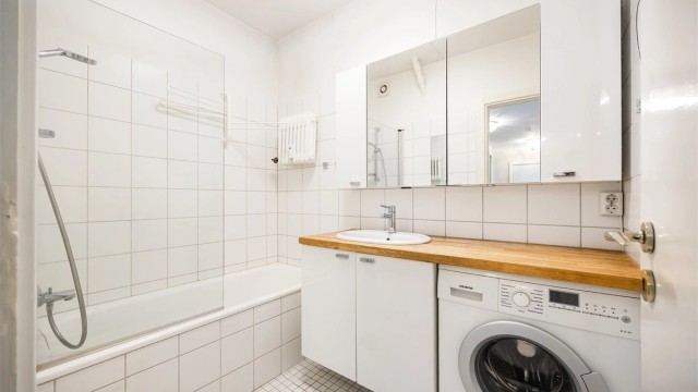 Lyst bad som er innredet med vegghengt servant med benkeplate i heltre og nedfelt vask. Innbygget badekar med glassvegg og dusjarmatur.