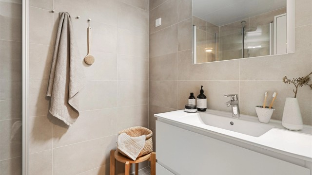 Stilrent baderom med opplegg for vaskemaskin, flislagt gulv med gulvvarme, flislagte vegger og metallplater i himling med downlights.