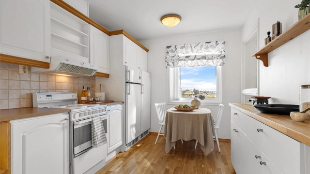 Kjøkkeninnredning fra byggeår med profilerte fronter og benkeplate med laminert overflate. Kjøkkenvask med armatur. Frittstående komfyr, oppvaskmaskin, kjøleskap og fryser. Ventilator integrert i overskap.