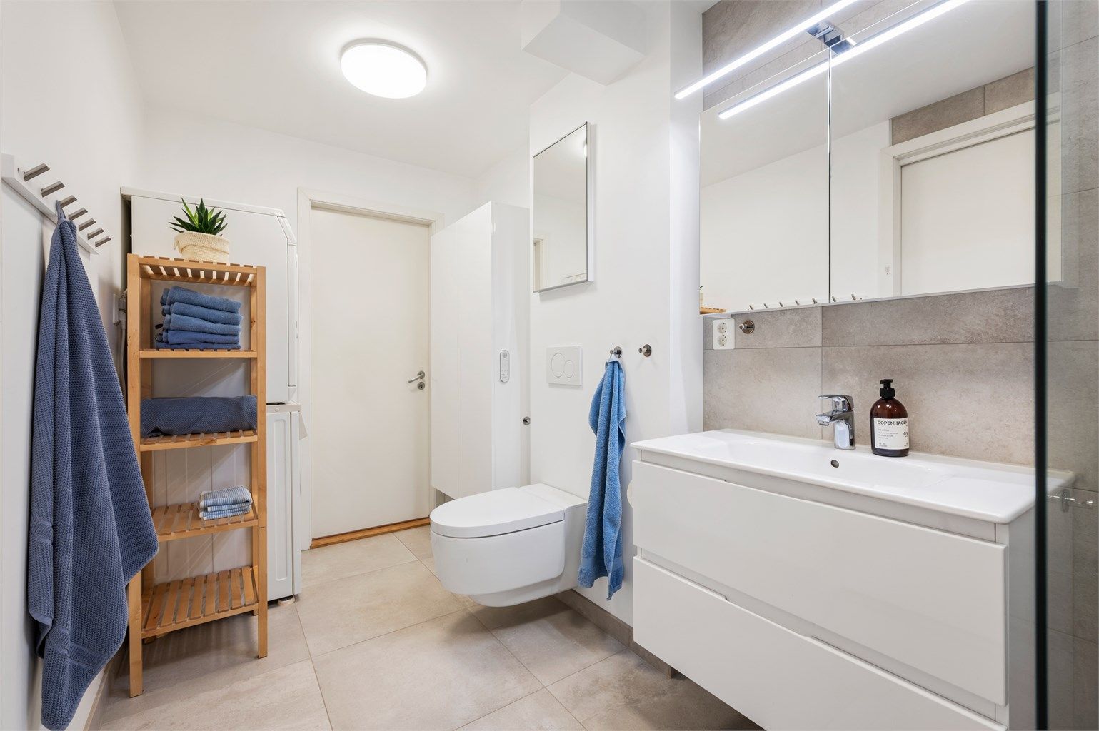 Tidløst flislagt bad med gulvvarme og opplegg for vaskemaskin. Videre er badet innredet med servantskap med speil med lys, vegghengt Geberit dusjttoalett og dusjhjørne. 