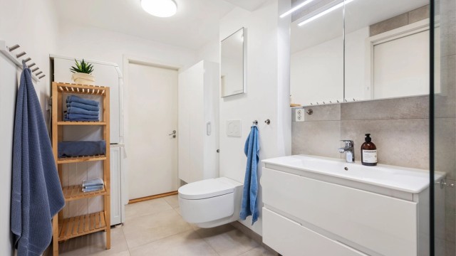 Tidløst flislagt bad med gulvvarme og opplegg for vaskemaskin. Videre er badet innredet med servantskap med speil med lys, vegghengt Geberit dusjttoalett og dusjhjørne.