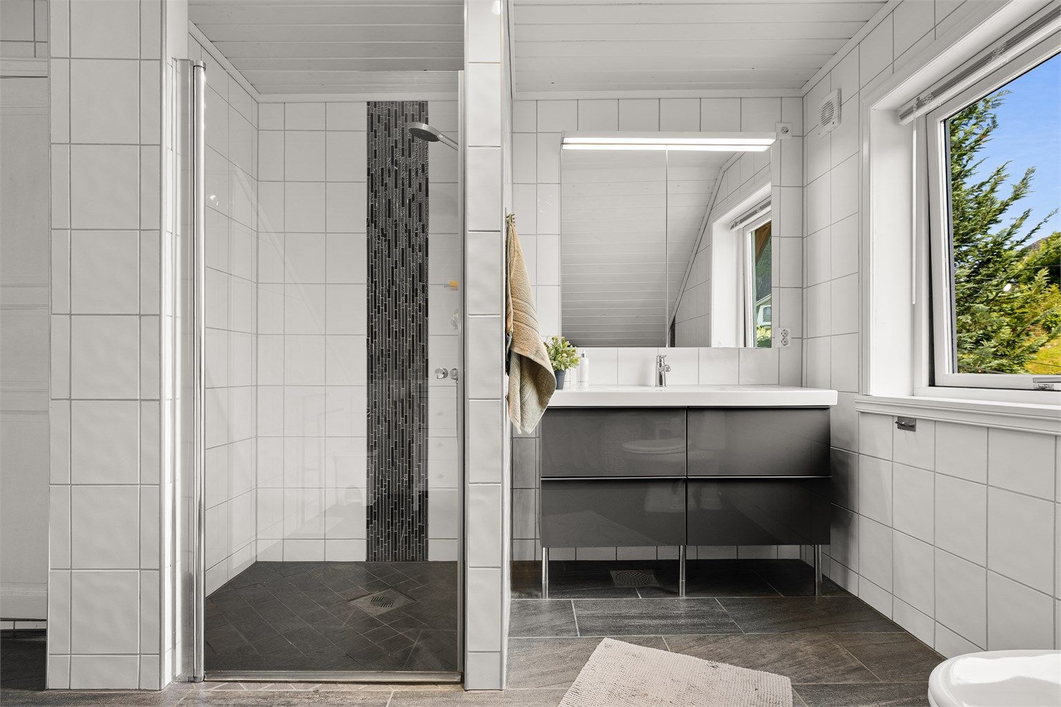 Badet er utstyrt med dusjhjørne, vegghengt servantinnredning og speilskap med overlys.