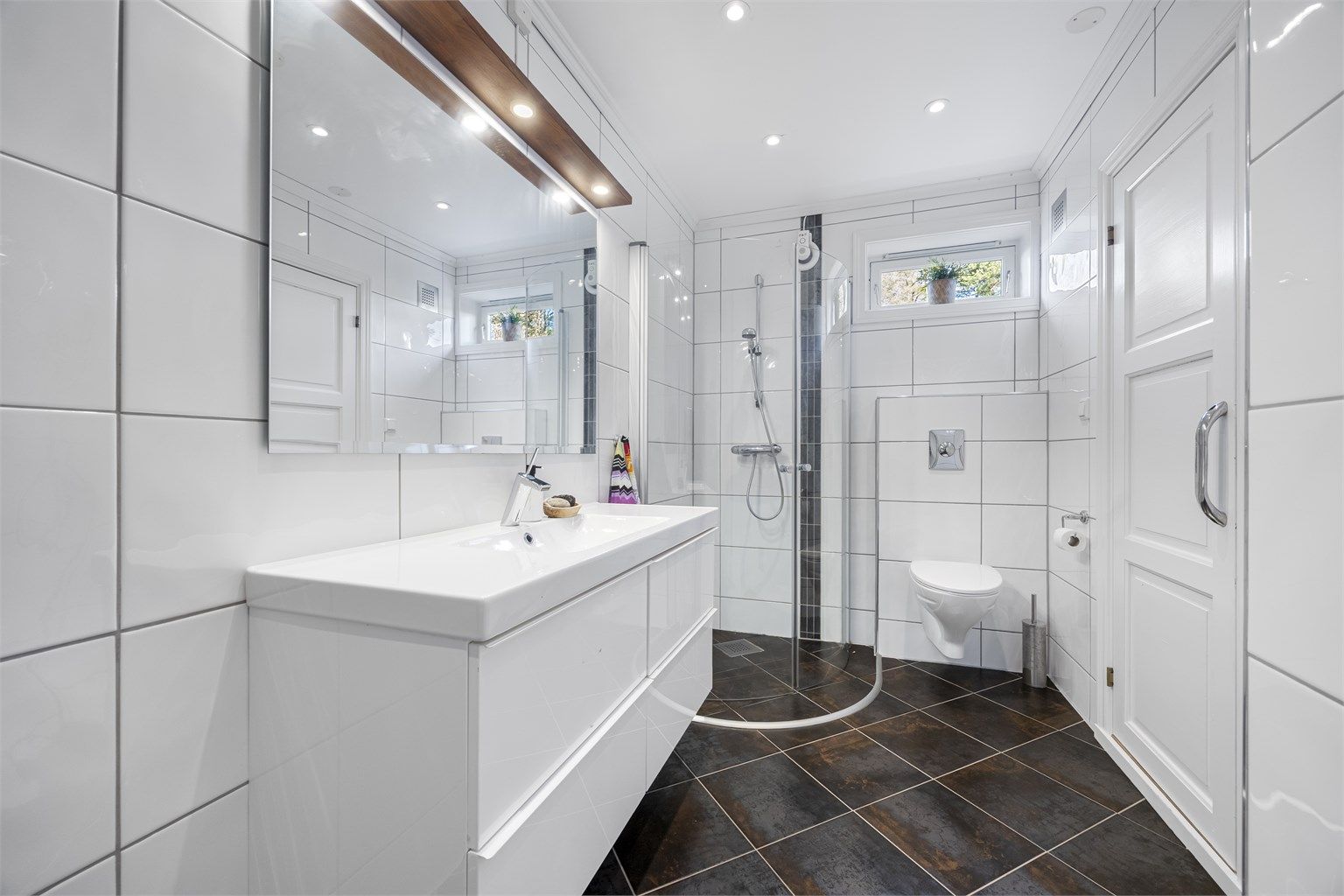 Badet i underetasjen har fliser på gulv og vegger, og er utstyrt med dusjvegger, toalett og en servant nedfelt i innredningen.