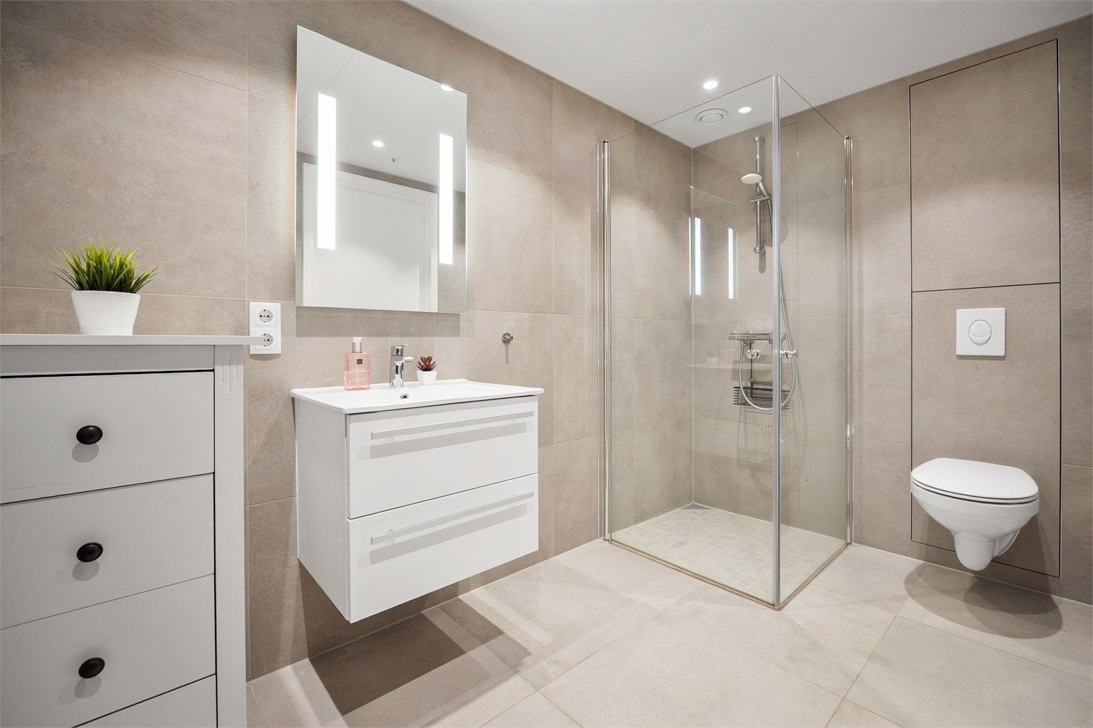 Stilrent bad 1 med store flisoverflater, ved entré. Badet har dusjhjørne med innfellbare dusjdører av glass.