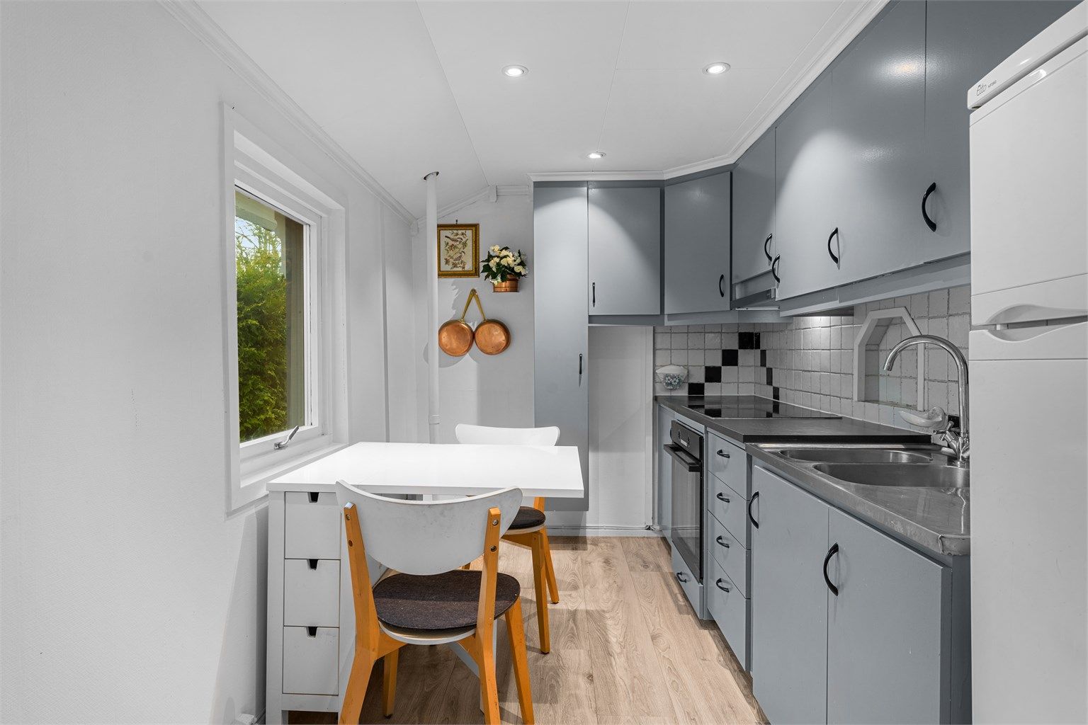 Eldre kjøkkeninnredning med glatte malte fronter, overskap og ventilator tilkoblet avtrekkskanal. Laminert benkeplate og heldekkende benkebeslag med skyllekum og oppvaskkum med to-greps armatur samt induksjons koketopp. 