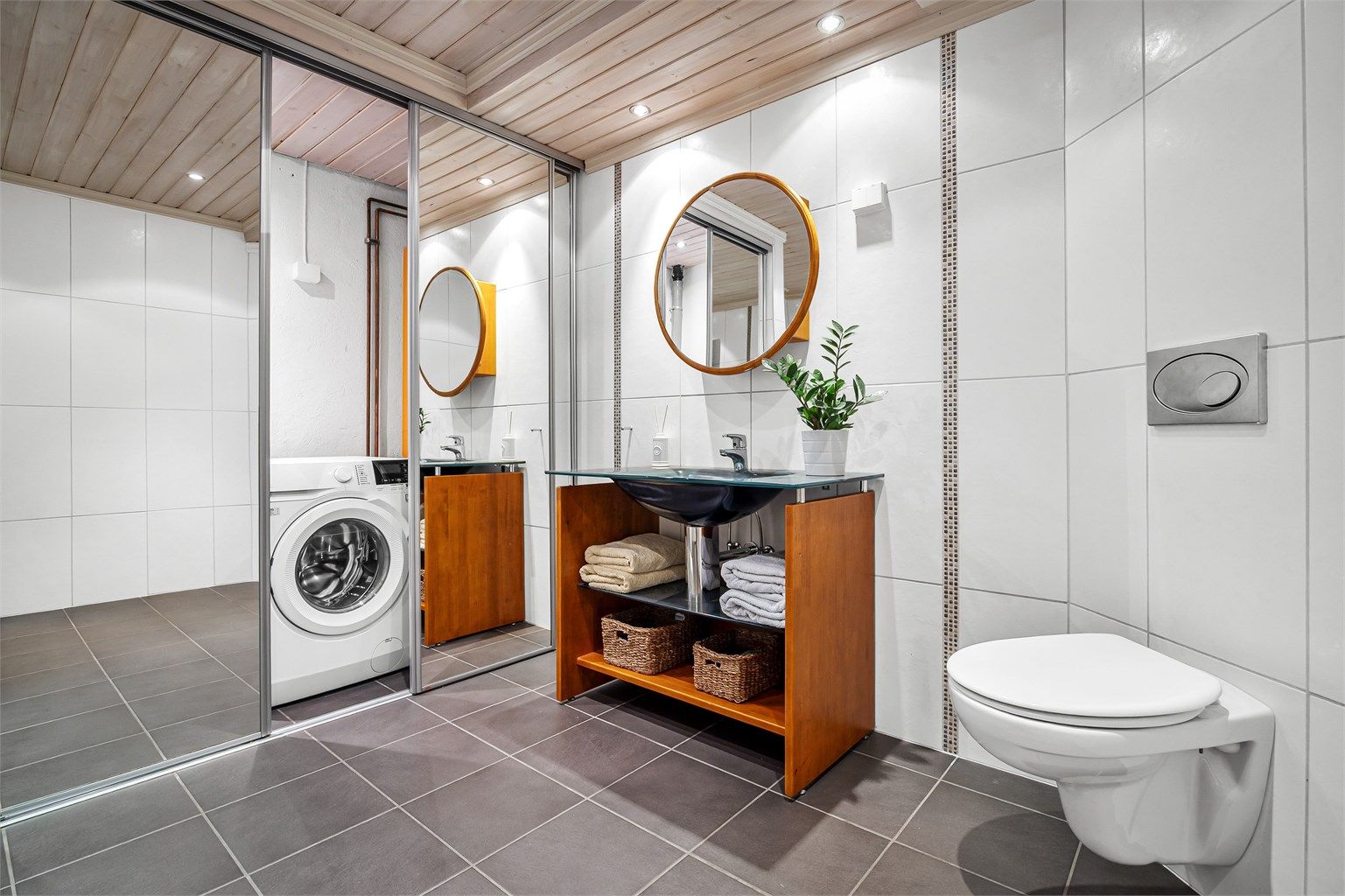 Badet er utstyrt med gulvstående innredning med heldekkende servant, badekar og vegghengt toalett. Bak skyvedør med speilfronter er det veggmontert utslagsvask, varmtvannsbereder og opplegg for vaskemaskin.