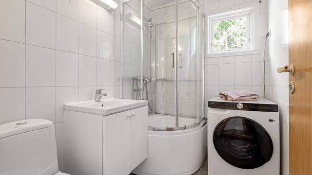Baderommet i første etasje er innredet med flislagt gulv, baderomsplater, dusjkabinett og opplegg for vaskemaskin.