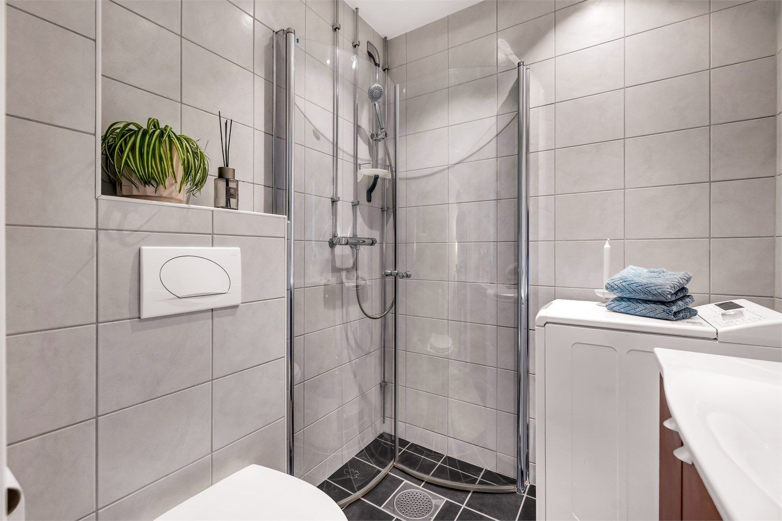 Dusjhjørne med innfellbare glassdører, vegghengt servant og toalett. Det er opplegg for vaskemaskin.