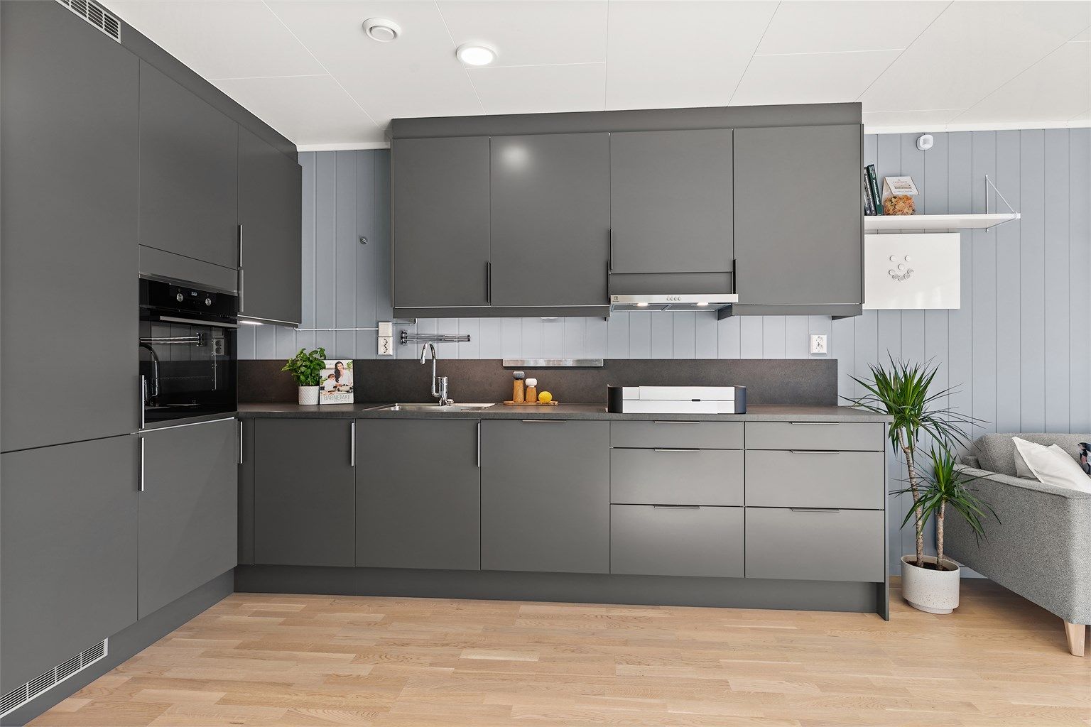 Stilrent og moderne kjøkken i praktisk L-form, med glatte mørkegrå fronter og en matchende veggplate som strekker seg elegant opp fra benkeplaten.
