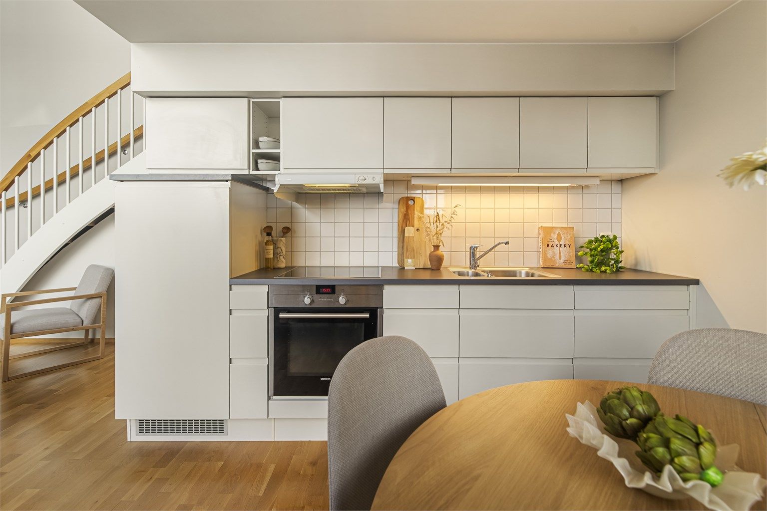 Det er montert en moderne kjøkkeninnredning med slette fronter og laminatbenkeplate. Fliser mellom benkeplate og overskap. Integrert oppvaskmaskin, platetopp, stekeovn og kjøleskap i høyskap medfølger.