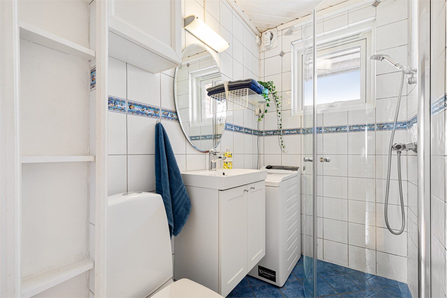 Flislagt baderom med gulvvarme innredet med gulvstående toalett, servant med underliggende skap, vegghengt speil med overbelysning og dusjhjørne. Nye dusjdører i 2026