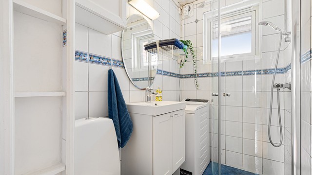 Flislagt baderom med gulvvarme innredet med gulvstående toalett, servant med underliggende skap, vegghengt speil med overbelysning og dusjhjørne. Nye dusjdører i 2026