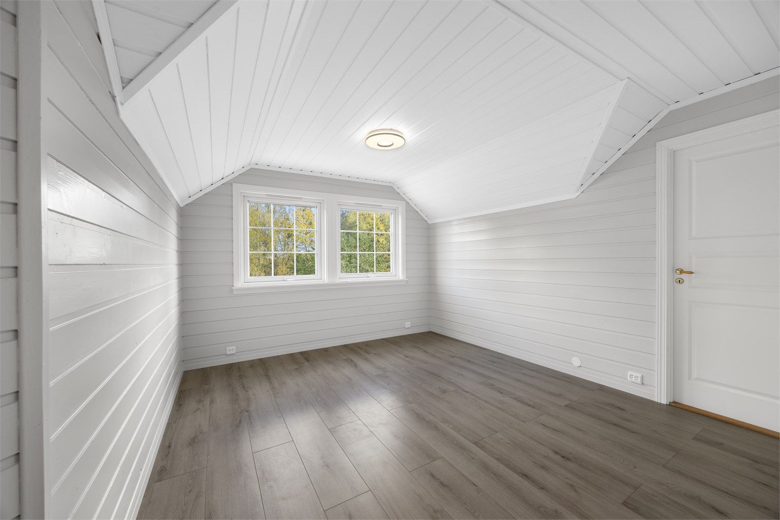 Loftstue med hvitmalt trepanel og vinduer mot hagen.