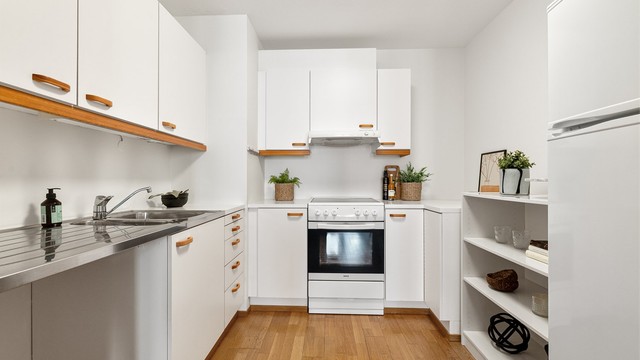 Kjøkkeninnredning med glatte, lyse fronter og benkeplater av laminat. Over komfyren er det ventilator med avtrekk, og husbrannslange er plassert under oppvaskkummen.
