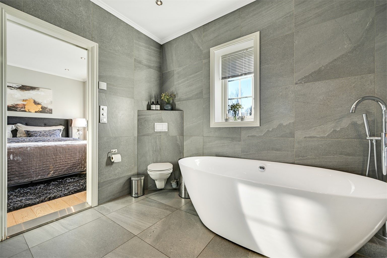 Badet er innredet med fliser på gulv og vegger, og har dobbel servant, veggmontert toalett, badekar samt dusjhjørne med glassvegger.