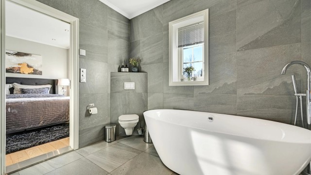 Badet er innredet med fliser på gulv og vegger, og har dobbel servant, veggmontert toalett, badekar samt dusjhjørne med glassvegger.