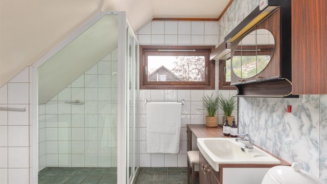 Badet er innredet med flislagt gulv med varme, delvis flislagte vegger kombinert med baderomsplater, samt dusjhjørne, servantinnredning og toalett. Rommet har fått en helheltsvurdering på TG3 iht. tilstandsrapporten.