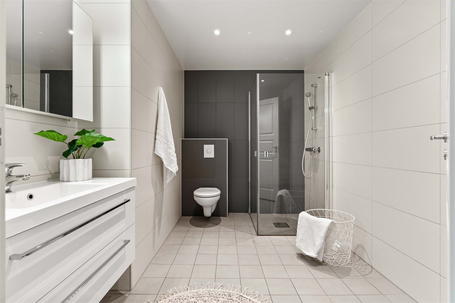 Stilrent, flislagt baderom med moderne innredning. Badet er utstyrt med vegghengt toalett, romslig servantmøbel med gode skuffer og lekkert dusjhjørne med innfellbare glassdører. Downlights i taket og nøytrale fargevalg skaper en behagelig atmosfære.