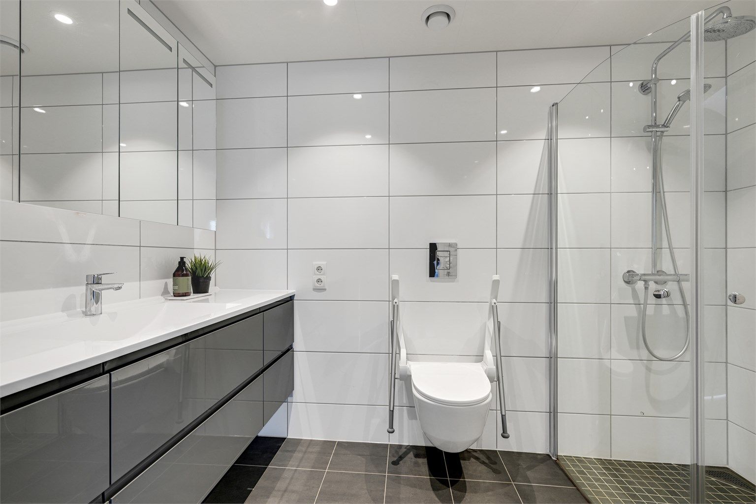Dette badet har fliser å gulv og vegger og varmekabler i gulvet. Badet inneholder servant i baderomsinnredningen, veggmontert toalett og dusjvegger i glass. 