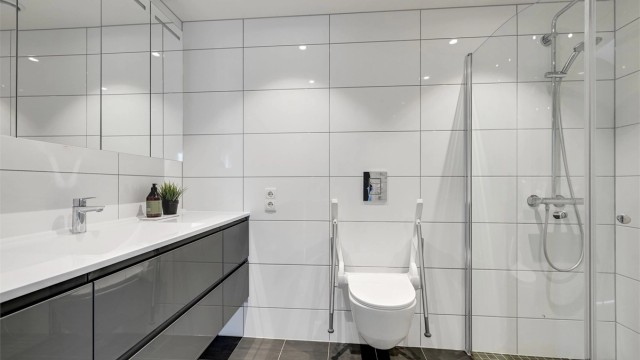 Dette badet har fliser å gulv og vegger og varmekabler i gulvet. Badet inneholder servant i baderomsinnredningen, veggmontert toalett og dusjvegger i glass.