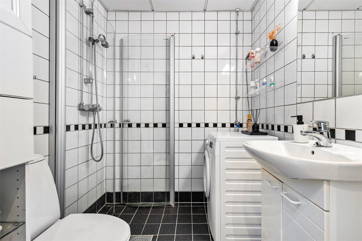 Badet er flislagt på gulv og vegger med elektrisk gulvvarme. Badet er innredet med dusjhjørne med svingbare glassdører, toalett og servant med innredning.