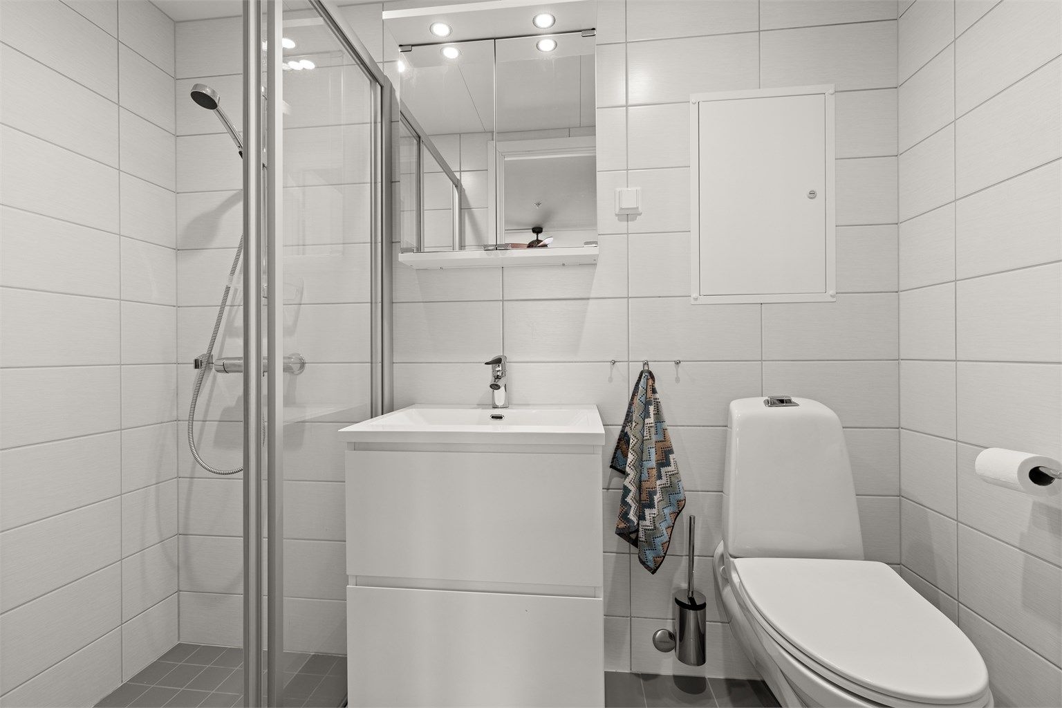 Badet har dusjhjørne, vegghengt toalett og speilskap.
