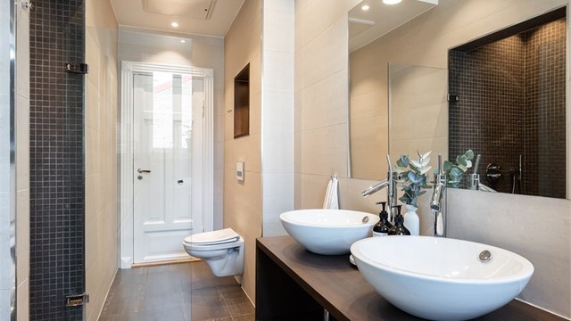 Badet er innredet med vegghengt toalett og servantinnredning med ovenpåliggende dobbelvask og speil med lys over vasken.