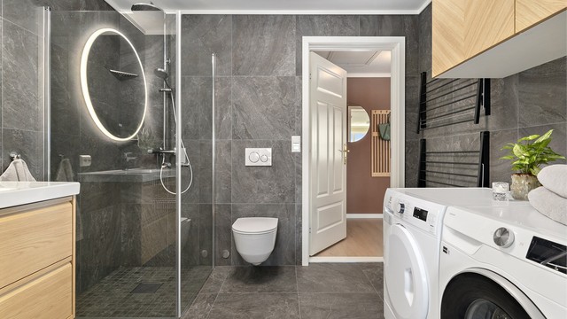 Badet har og opplegg for vaskemaskin og tørketrommel, vegghengt toalett, gulvvarme og praktisk løsning for tørking av klær. Legg og merke til overskap, som gir ekstra oppbevaringsplass