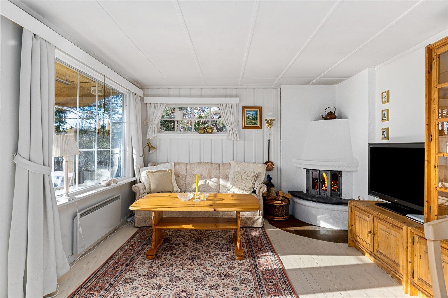 HYGGESTUEN: Peisen er plassert i hjørne slik at flammene kan sees fra hele rommet. Med malte flater og stort vindu mot vest er rommet lyst og trivelig.