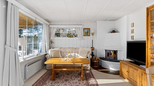 HYGGESTUEN: Peisen er plassert i hjørne slik at flammene kan sees fra hele rommet. Med malte flater og stort vindu mot vest er rommet lyst og trivelig.