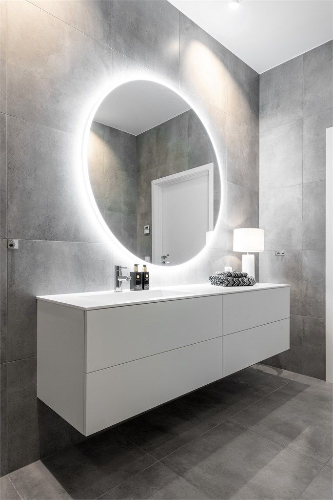 Stor romslig baderomsinnredning fra Azzurro med hvite glatte fronter og nedfelt vask med ett-greps armatur. Rundt speil med lys og stikkontakter over servant. 