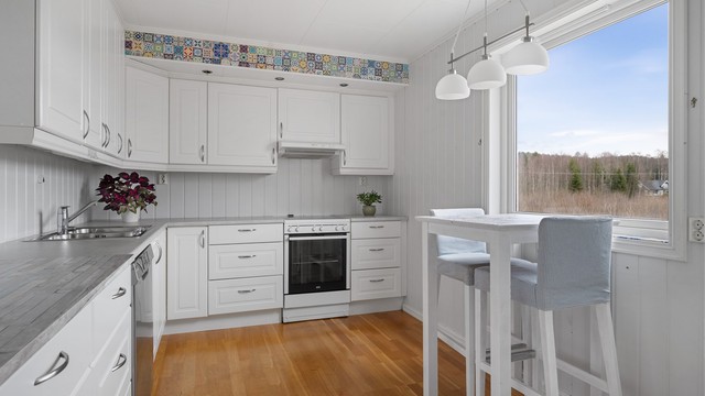 Kjøkkeninnredningen har hvite, profilerte fronter, laminert benkeplate og en praktisk frokostplass ved vinduet.