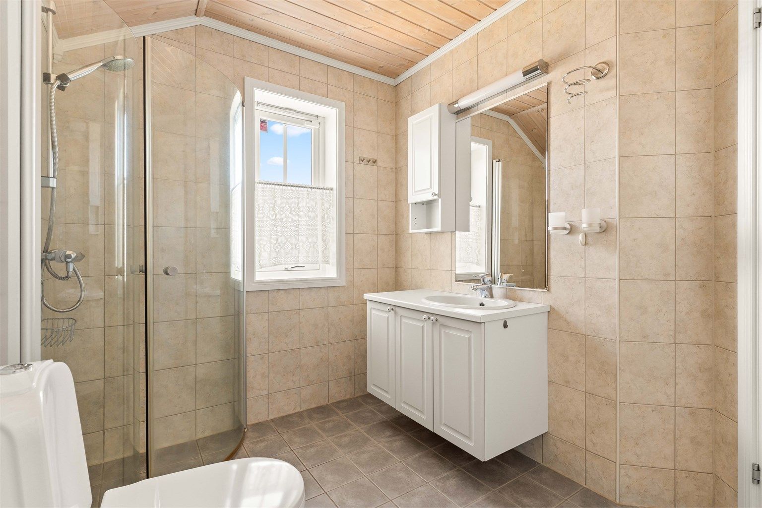 Badet er utstyrt med dusjnisje, gulvmontert toalett og servant med speil og overlys.