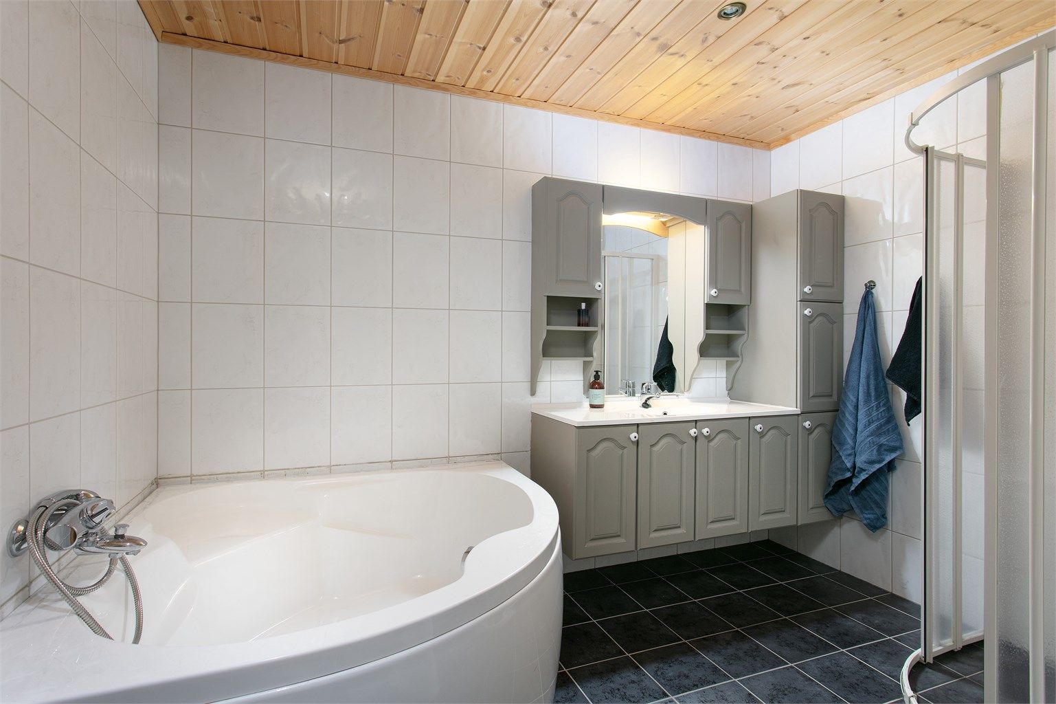 Badet har flislagt gulv med varmekabler, hjørnebadekar, dusjhjørne og grå baderomsinnredning.